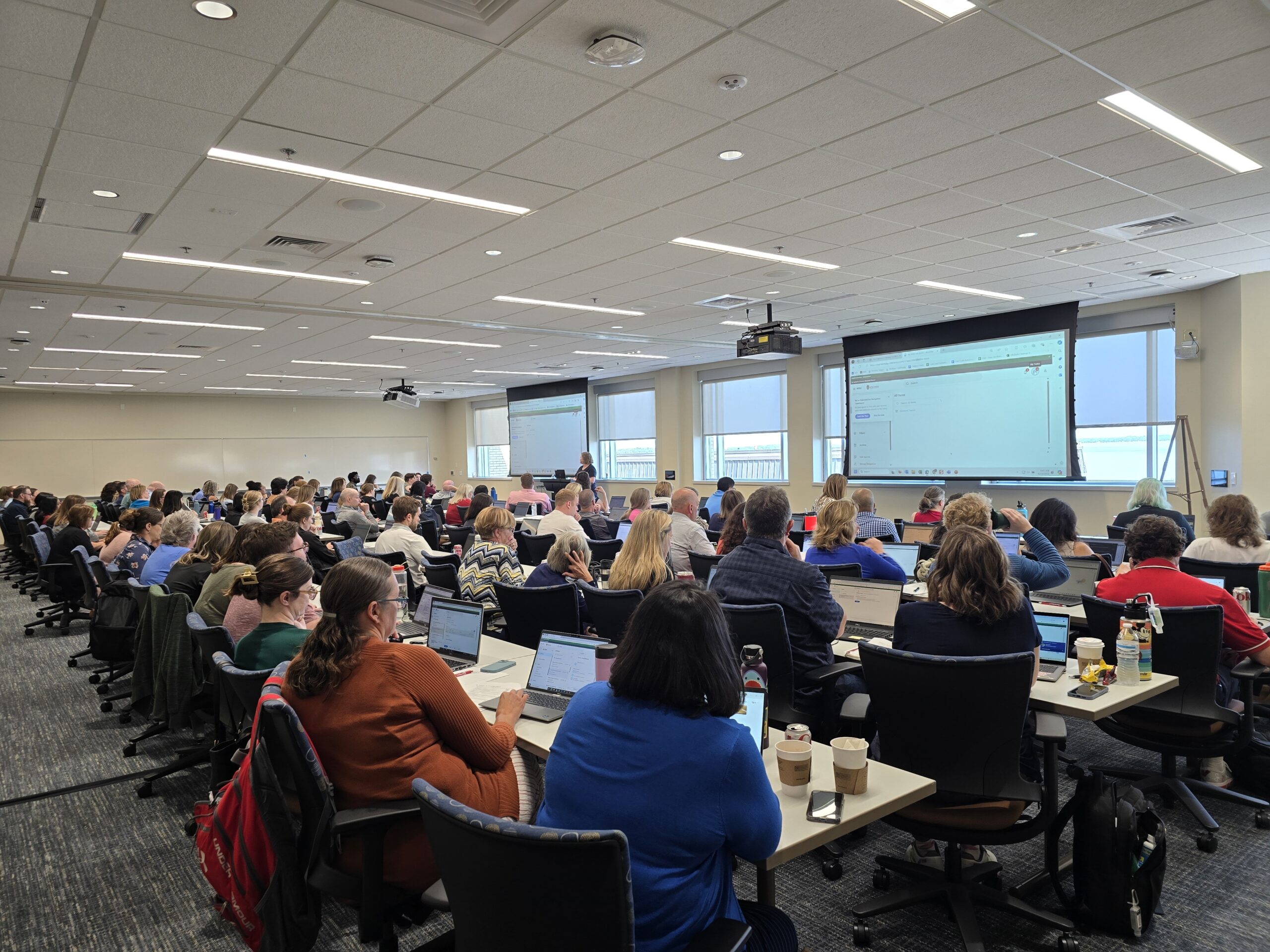 Dozens of staff sit behind computers in a large conference room with two projection screens at the front.