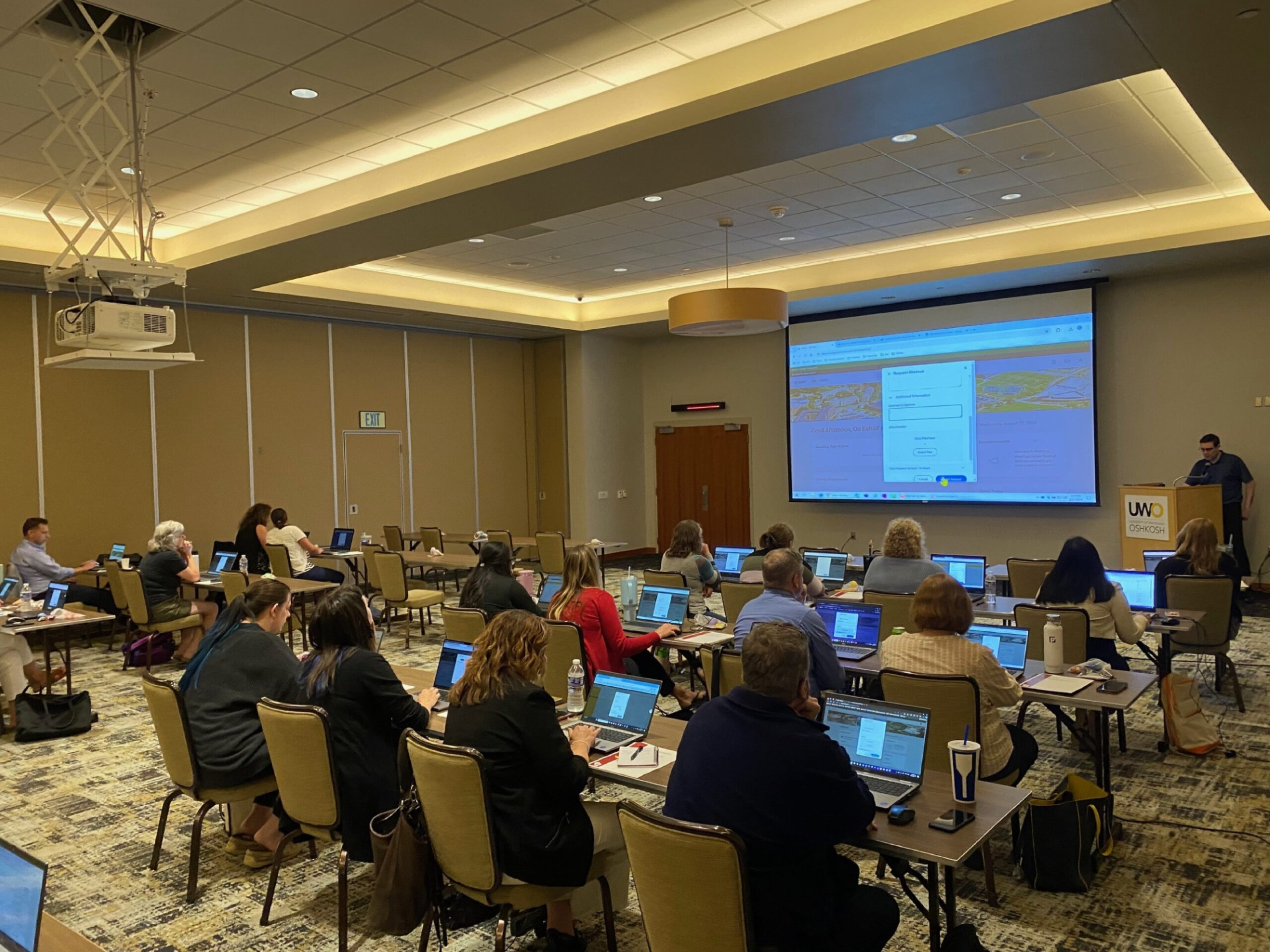 A large conference room is filled with people typing on laptops and facing a projection screen depicting the Workday interface.