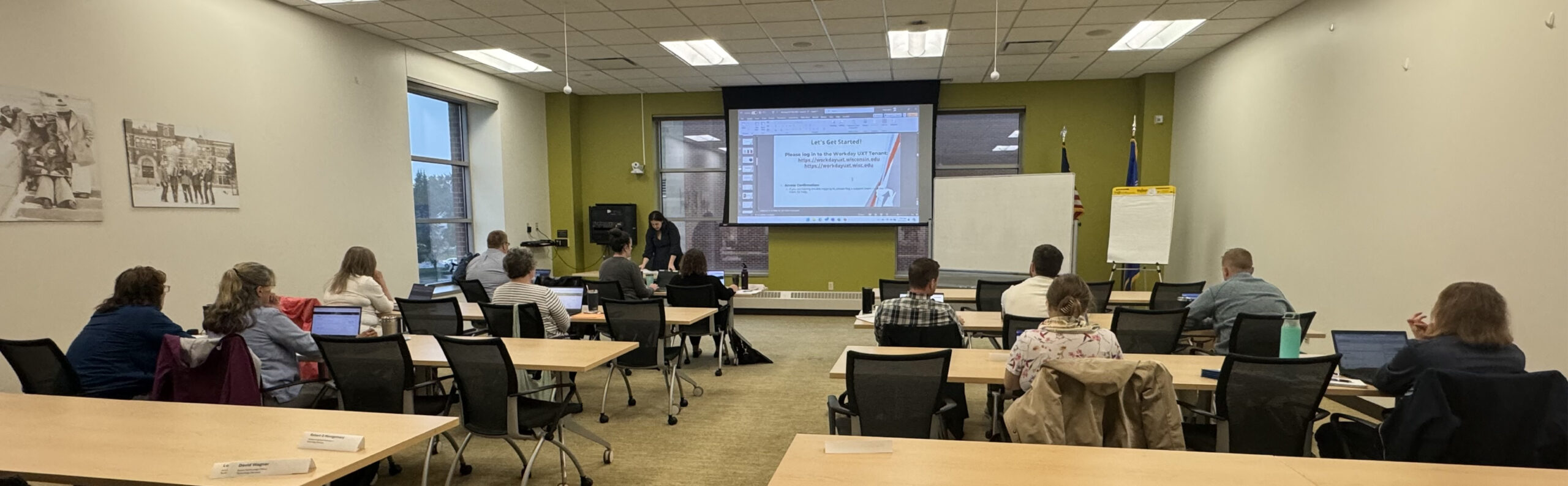 A dozen staff sit behind computers in a conference room with a projection screen and presenter at the front.